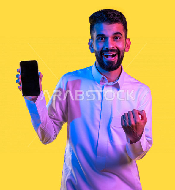 بورتريه لشاب عربي خليجي سعودي يقوم بعرض جهاز الهاتف المحمول بإيماءات تدل على السعادة و الفوز ، الحصول على الهدف المراد ، التسويق الرقمي ، اعلانات تسويقية ترويجية ، تطبيقات الهاتف الذكي ، خلفية صفراء