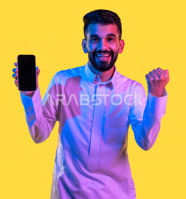 بورتريه لشاب عربي خليجي سعودي يقوم بعرض جهاز الهاتف المحمول بإيماءات تدل على السعادة و الفوز ، الحصول على الهدف المراد ، التسويق الرقمي ، اعلانات تسويقية ترويجية ، تطبيقات الهاتف الذكي ، خلفية صفراء