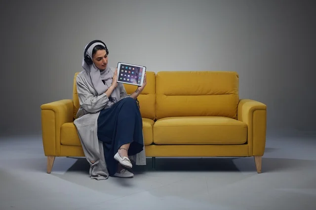 امرأة سعودية تحمل جهازاً لوحياً على أريكة صفراء