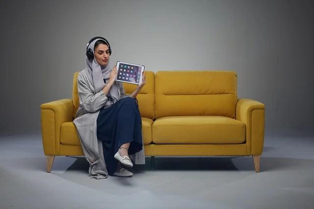 امرأة سعودية تحمل جهاز لوحي وسماعات على أريكة صفراء