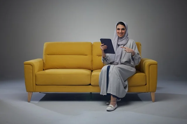 امرأة سعودية تستخدم جهازاً لوحياً على أريكة صفراء