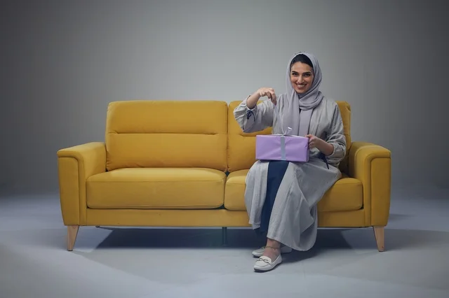 امرأة سعودية تجلس على أريكة صفراء تحمل علبة هدايا مغلفة