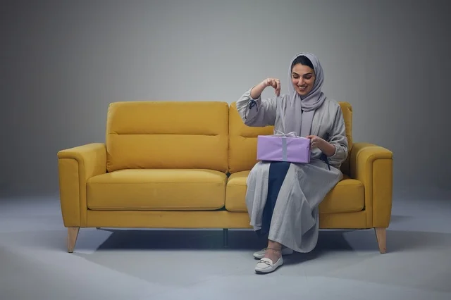 امرأة سعودية تفتح صندوق هدايا على أريكة صفراء في استوديو