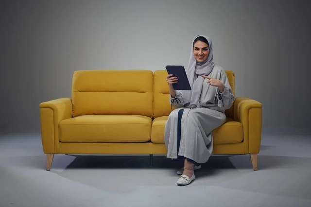 امرأة سعودية على أريكة صفراء تشير إلى جهاز لوحي