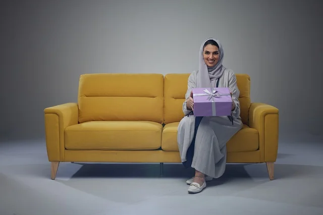 امرأة سعودية تجلس على أريكة صفراء تحمل علبة هدايا