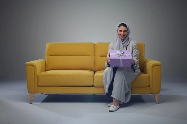 امرأة سعودية تجلس على أريكة صفراء تحمل صندوق هدايا امرأة سعودية تجلس على أريكة صفراء تحمل صندوق هدايا