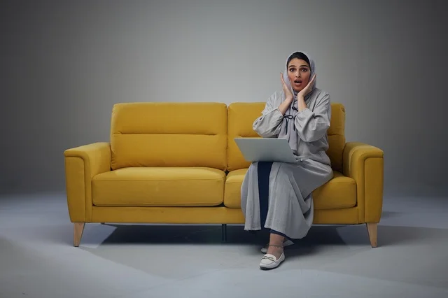 امرأة سعودية مندهشة مع حاسوب محمول على أريكة صفراء