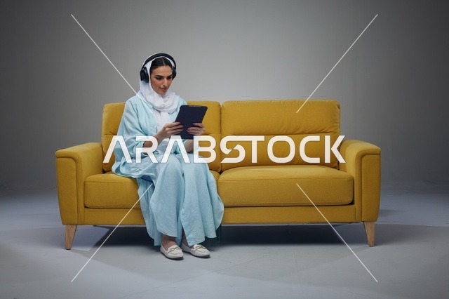 امرأة سعودية تستخدم جهاز لوحي وسماعات على أريكة صفراء