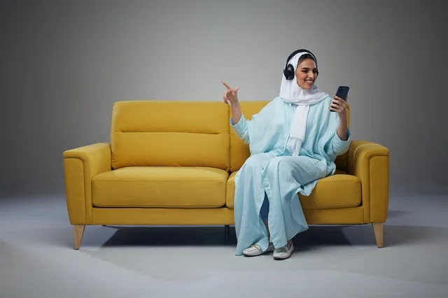 امرأة سعودية على أريكة صفراء مع هاتف ذكي وسماعات