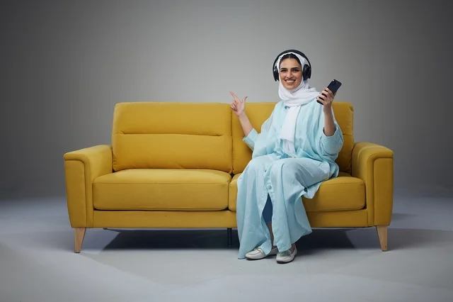 امرأة سعودية على أريكة صفراء مع سماعات وهاتف