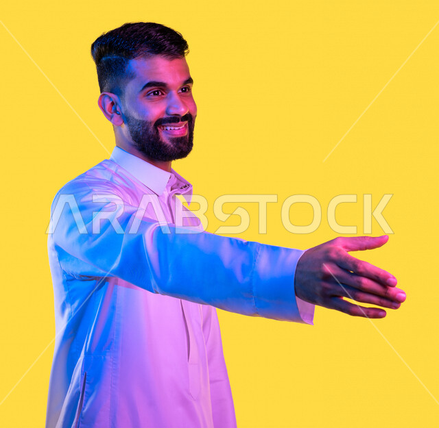 بورتريه لشاب عربي خليجي سعودي يمد يده للمصافحة بإيماءات و تعابير وجه تدل على السعادة ، التسليم و المصافحة باليد ، التفاعل الاجتماعي و الحياة المجتمعية ، خلفية صفراء
