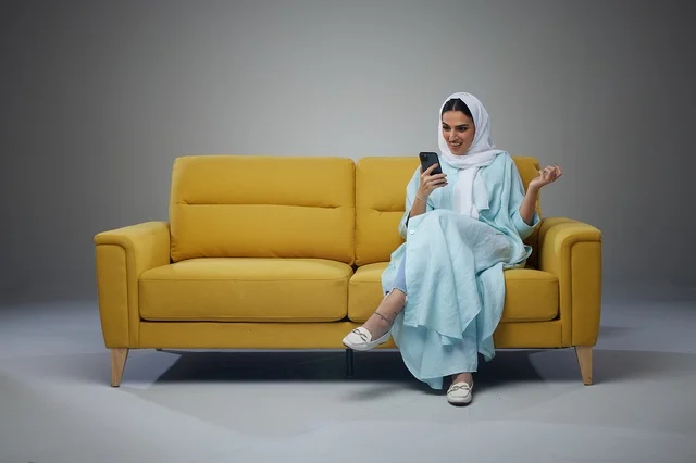 امرأة سعودية بعباءة تستخدم الهاتف الذكي على أريكة صفراء
