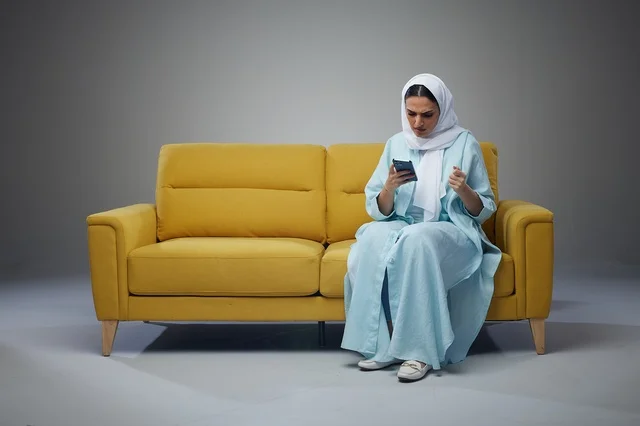 امرأة سعودية تجلس على أريكة صفراء تستخدم الهاتف الذكي