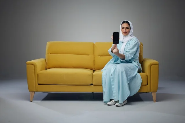 امرأة سعودية تجلس على أريكة صفراء وتستخدم هاتف ذكي
