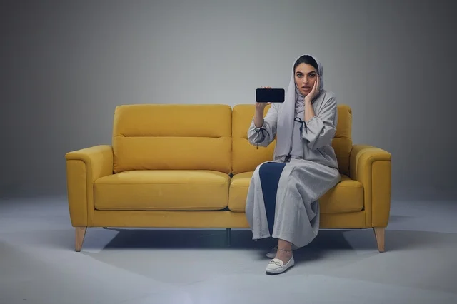 Saudi Woman on Yellow Sofa Holding Smartphone Surprised