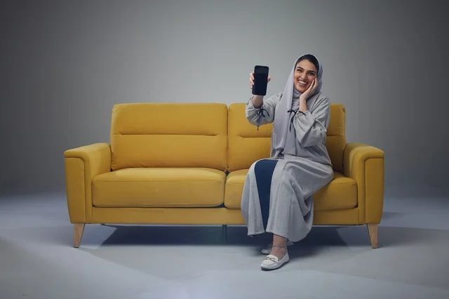 امرأة سعودية تحمل هاتفاً ذكياً على أريكة صفراء في الاستوديو