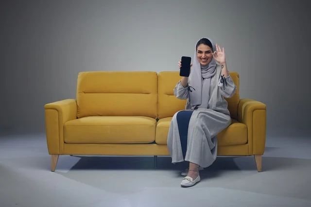 امرأة سعودية تحمل هاتف ذكي على أريكة صفراء في استوديو