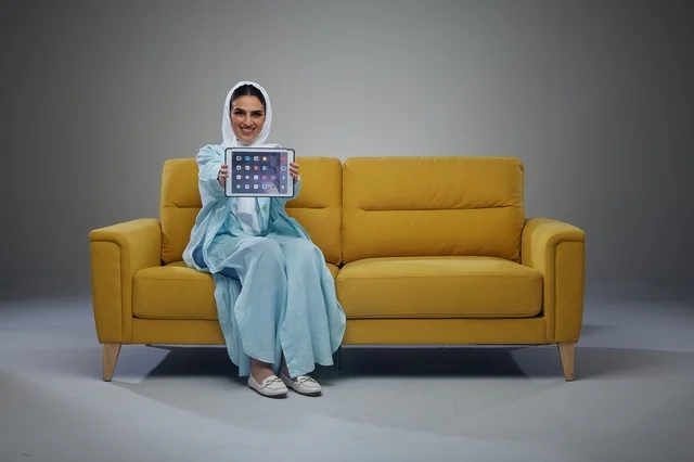 امرأة سعودية على أريكة صفراء تحمل جهازاً لوحياً