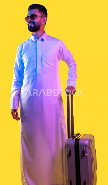 Portrait of a Saudi Arabian Gulf youth ready to travel, wearing sunglasses and carrying a travel bag, watching something on the tour, spending a fun leisure trip, convalescence and travel, yellow background