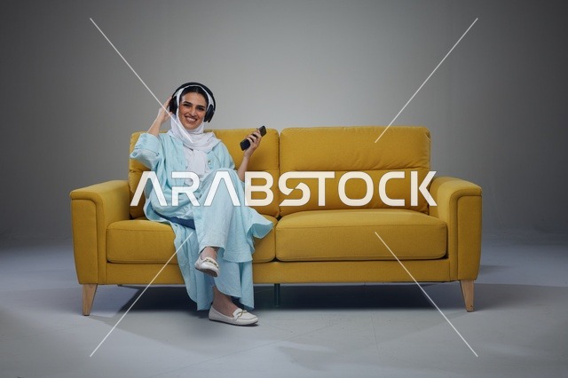 امرأة سعودية تستخدم هاتف ذكي وسماعات على أريكة صفراء