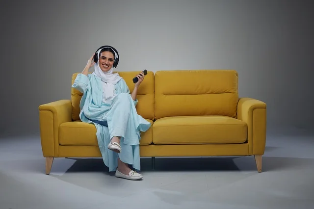 Saudi Woman Using Smartphone with Headphones on Yellow Sofa