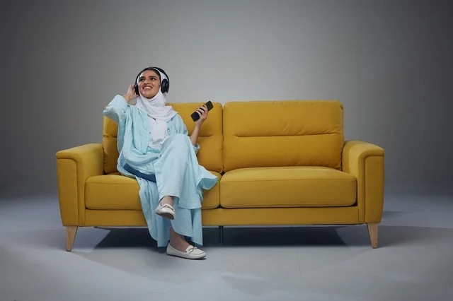 امرأة سعودية على أريكة صفراء تستخدم سماعات الرأس