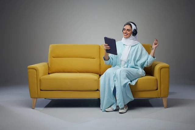 Saudi Woman Using Tablet with Headphones on Yellow Sofa