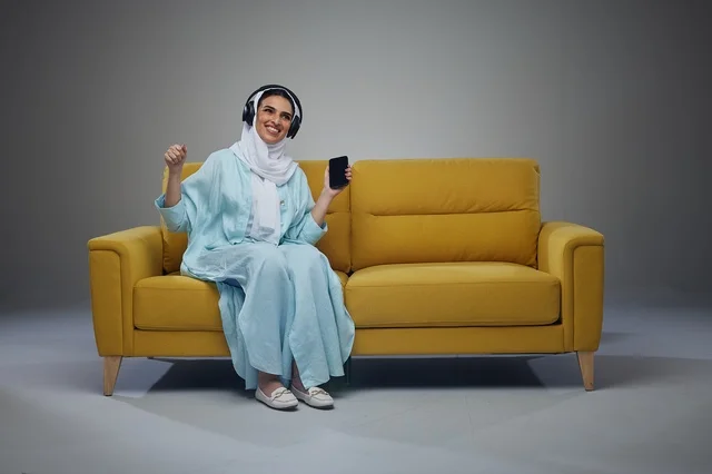 امرأة سعودية على أريكة صفراء تستخدم الهاتف والسماعات
