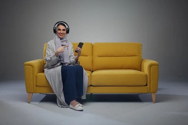 امرأة سعودية على أريكة صفراء مع هاتف ذكي وسماعات