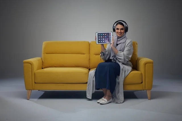 امرأة سعودية تجلس على أريكة تستخدم جهازًا لوحيًا مع سماعات