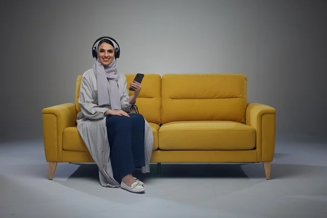 امرأة سعودية على أريكة صفراء مع هاتف ذكي وسماعات