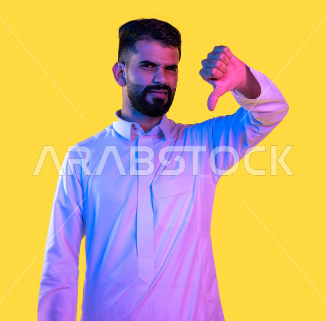 بورتريه لشاب عربي خليجي سعودي يشير بأصبع الابهام لأسفل بإيماءات وتعابير وجه و يدين تدل على الفشل و الرفض ، عدم الموافقة و الرضا ، خلفية صفراء