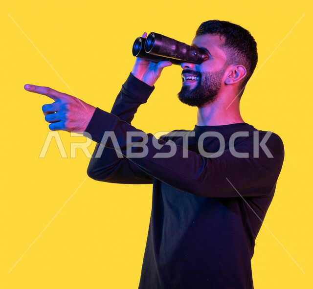 بورتريه لشاب عربي خليجي سعودي يمسك بيده ناظور و يشير بيده تجاه شئ ما رآه ، مشاهدة شيئا ما بإيماءات و تعابير وجه و يدين تدل على السعادة ، رؤية مستقبلية ، التخطيط المستقبلي ، أهداف و طموحات ، خلفية صفراء