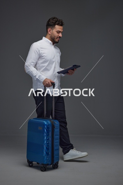 مسافر سعودي يستخدم جهازًا لوحيًا مع حقيبة سفر وسماعات