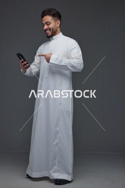 رجل سعودي يرتدي الثوب التقليدي ويشير إلى هاتف ذكي