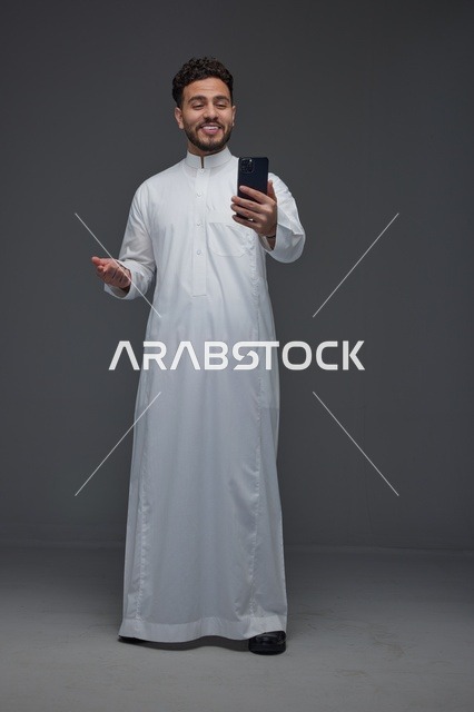 رجل سعودي مبتسم بالثوب التقليدي يستخدم الهاتف الذكي