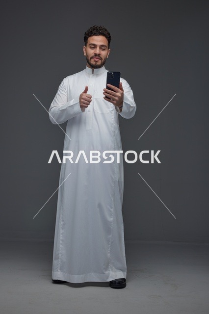 رجل سعودي بالثوب يستخدم الهاتف الذكي مع إشارة الإبهام