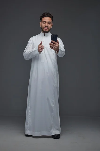 رجل سعودي بالثوب يستخدم الهاتف الذكي مع إشارة الإبهام
