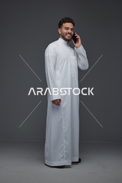 رجل سعودي يبتسم يتحدث بالهاتف الذكي بالثوب التقليدي