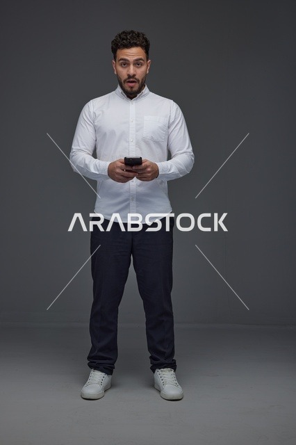 رجل سعودي مندهش يستخدم الهاتف الذكي بخلفية رمادية