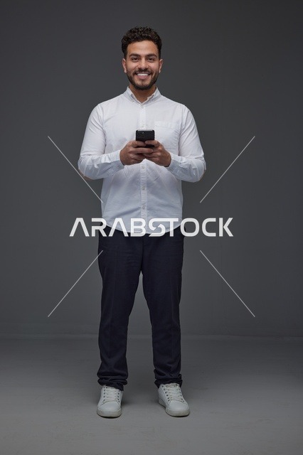 رجل سعودي مبتسم يستخدم الهاتف الذكي في استوديو رمادي