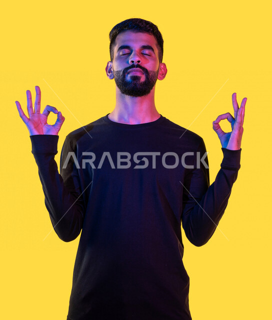 بورتريه لشاب عربي خليجي سعودي يغمض عينيه و يرفع يديه إلى أعلى بإيماءات و تعابير وجه و يدين تدل التأمل و الاسترخاء ، ممارسة تمارين رياضة اليوغا ، تمرين التنفس العميق ، خلفية صفراء