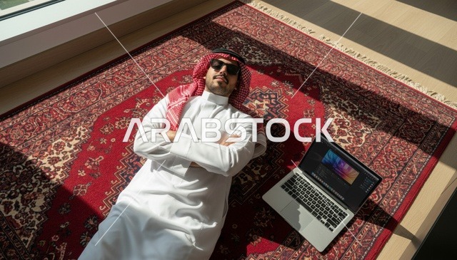رجل سعودي بالثوب التقليدي يستلقي على سجادة مع لابتوب