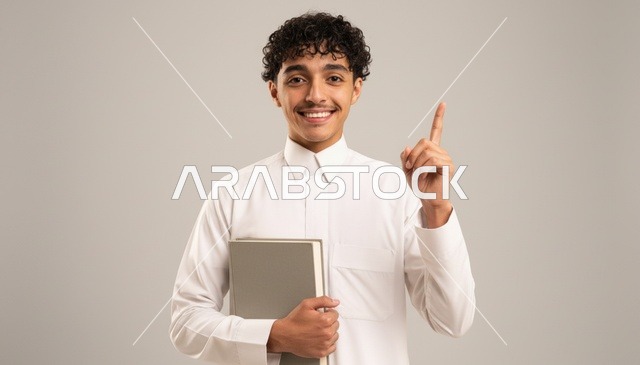 شاب سعودي بالثوب التقليدي يحمل كتاباً ويشير للأعلى
