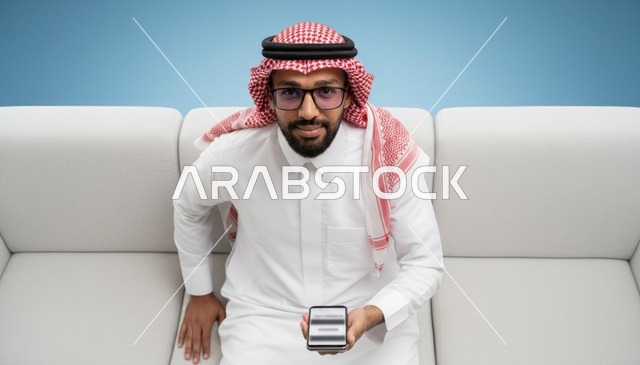 رجل سعودي بالزي التقليدي يمسك هاتف ذكي على الأريكة