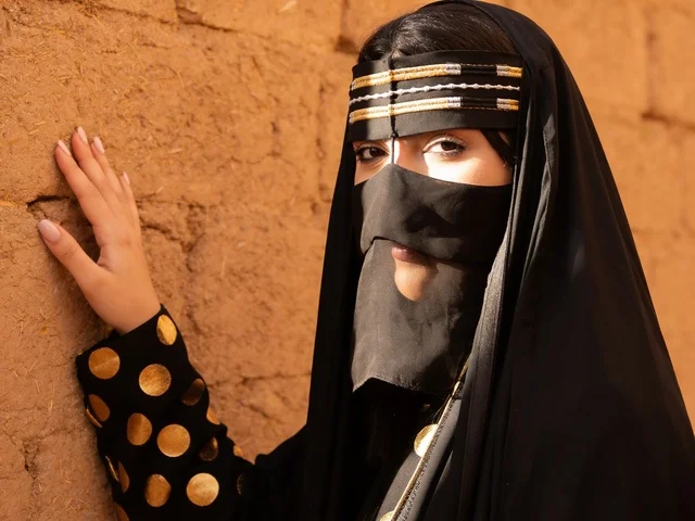 امرأة سعودية بالزي النجدي التقليدي أمام جدار طيني