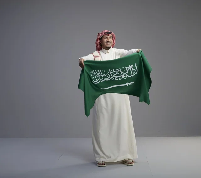 رجل سعودي بزي تقليدي يحمل العلم الوطني في استوديو رمادي
