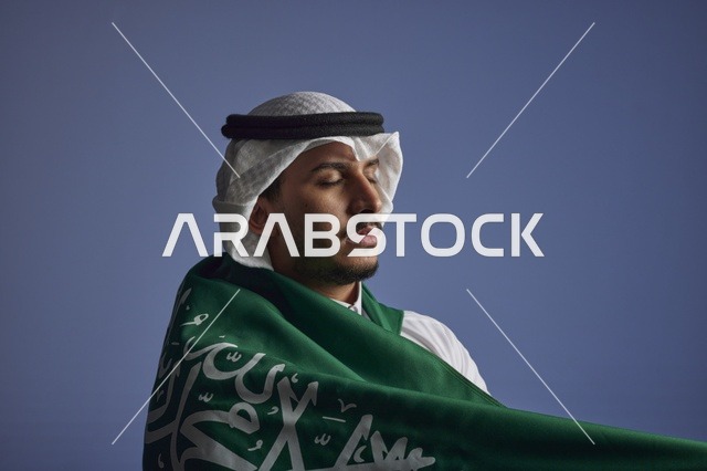 شاب سعودي يلتف بالعلم السعودي بخلفية زرقاء