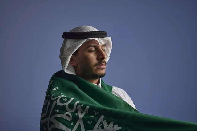 شاب سعودي يلتف بالعلم السعودي بخلفية زرقاء