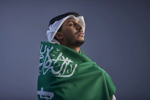 شاب سعودي يلتف بالعلم السعودي وخلفية رمادية